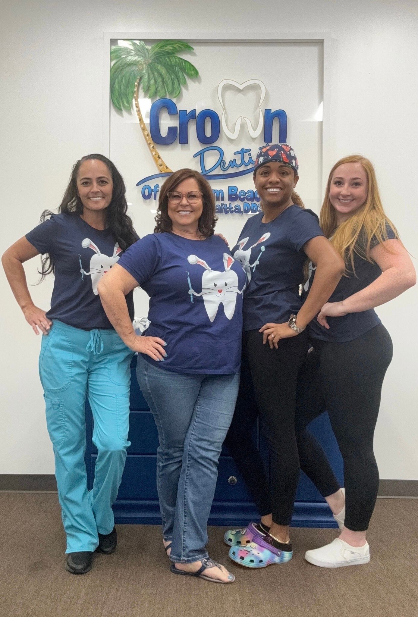 Four dental office staff members pose in front of a sign. They wear matching blue shirts, some in scrubs, smiling with hands on hips.