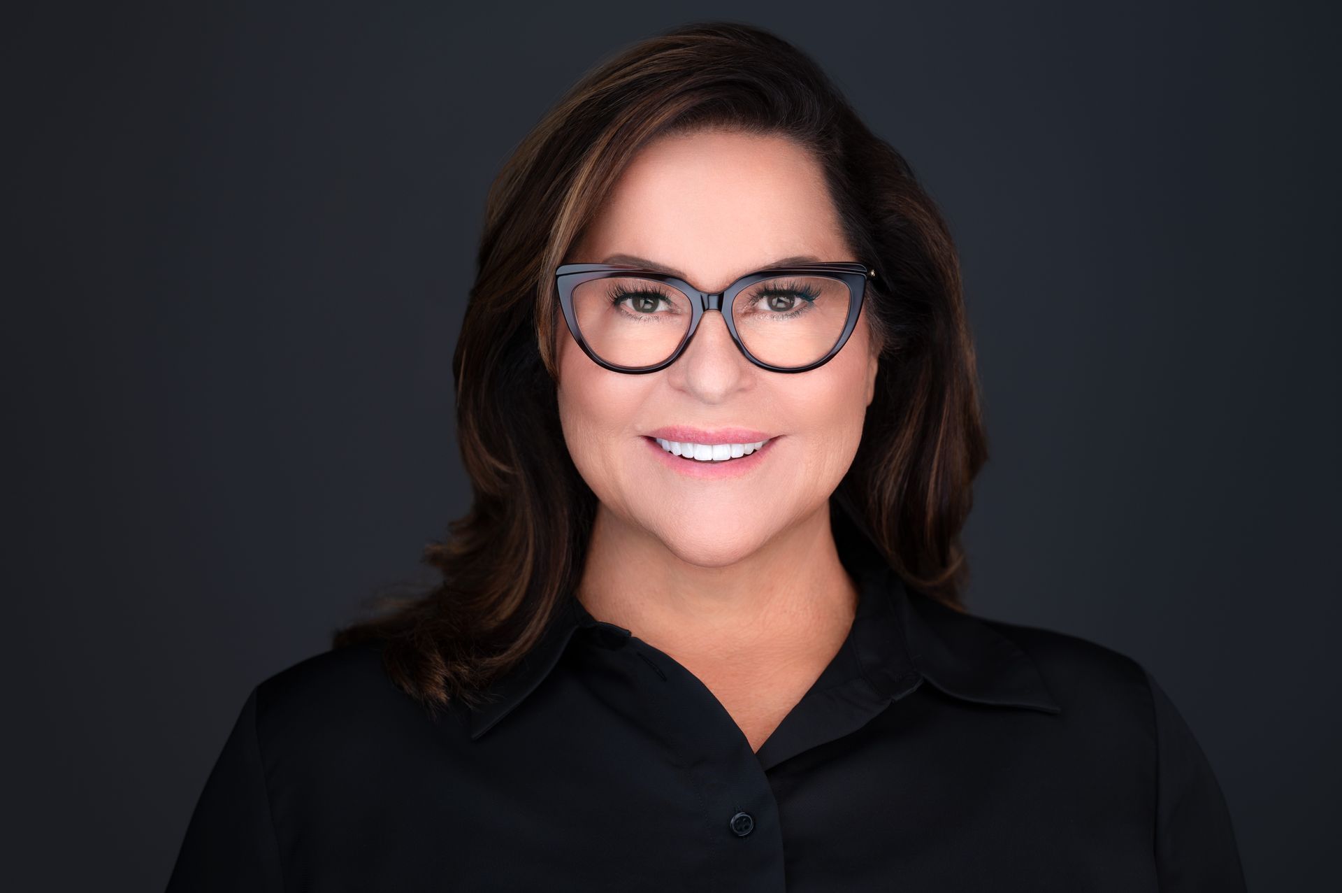 Woman with glasses, smiling, wearing a black shirt, against a dark gray backdrop.