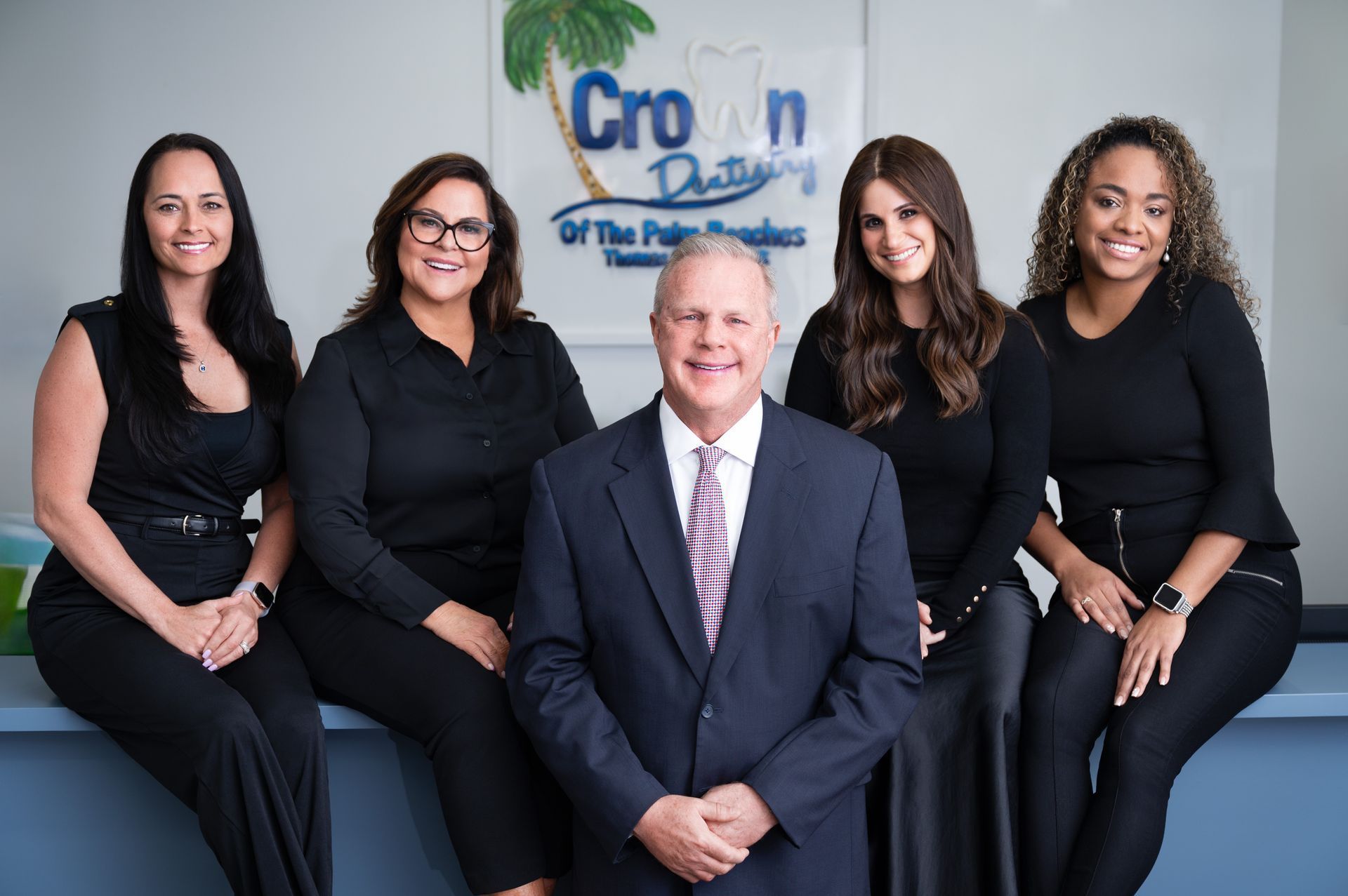 Staff of Crown Dentistry posing together. A man in a suit is centered, surrounded by four women in black. Palm tree logo backdrop.
