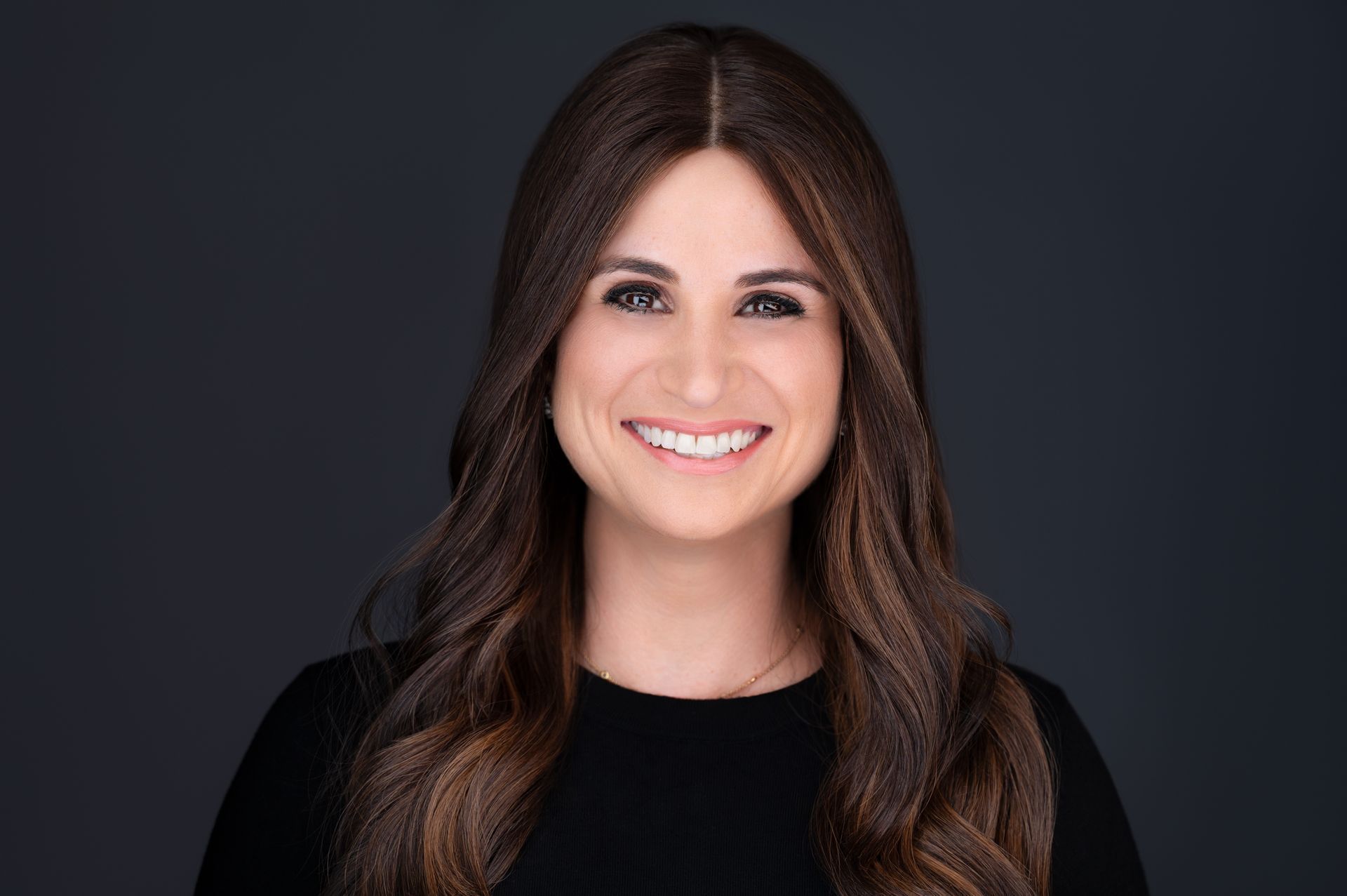 Woman with long, brown hair smiling at the camera, wearing a black top, against a dark gray background.