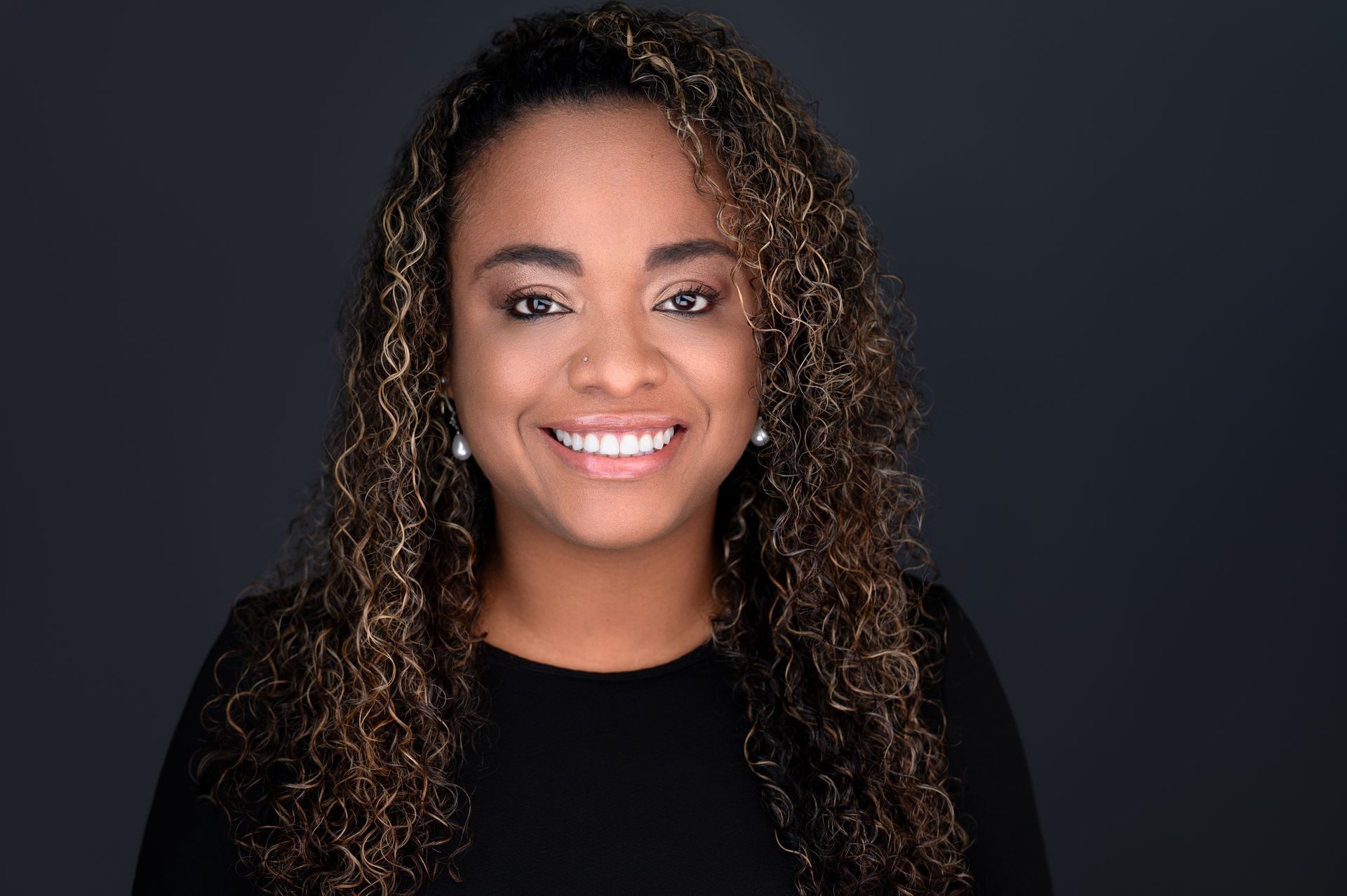 Woman with curly hair smiles, wearing a black top, against a dark gray background.