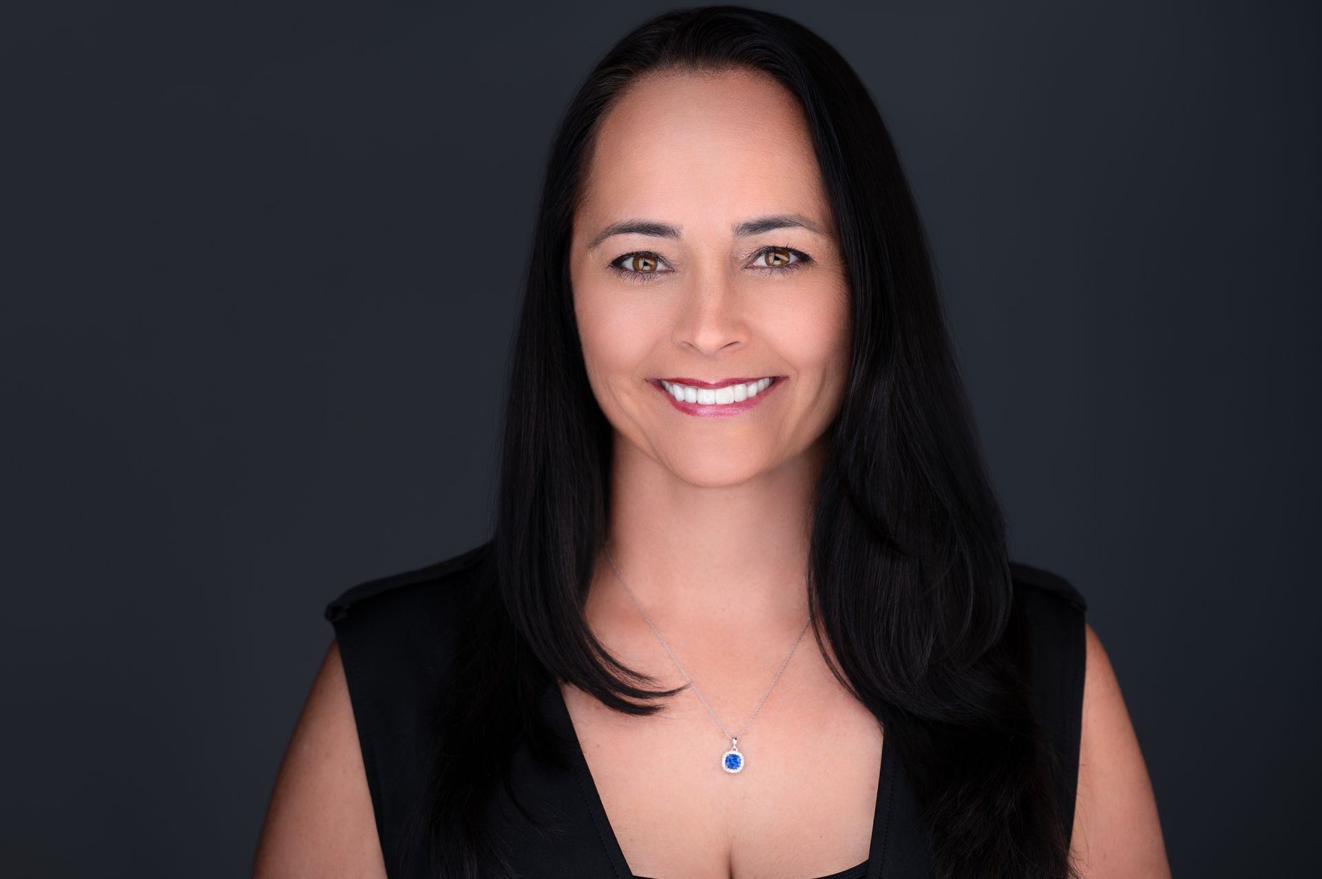 Woman with dark hair smiles, wearing a black top and a necklace against a dark gray background.