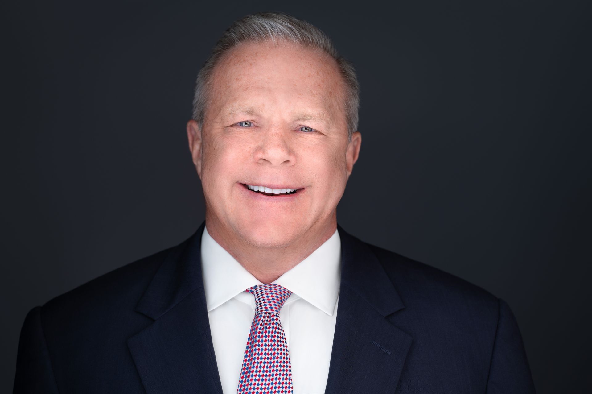 Smiling man in a suit, white shirt, and patterned purple tie, against a dark gray background.