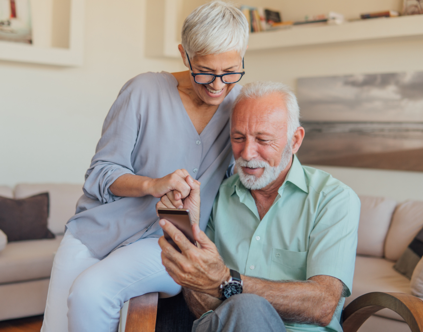 Elderly couple looking at a smartphone, woman points, both smiling, indoor setting.
