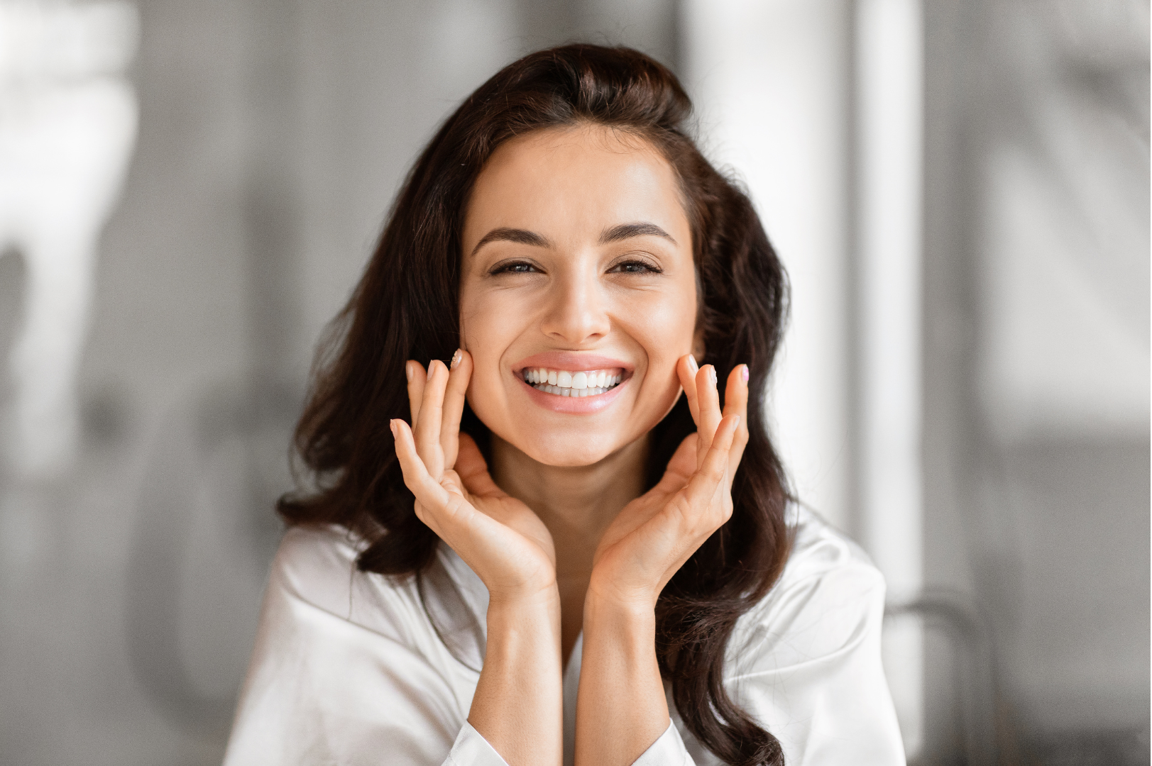 Woman with long brown hair smiles while touching her cheeks. Bright, indoor setting.