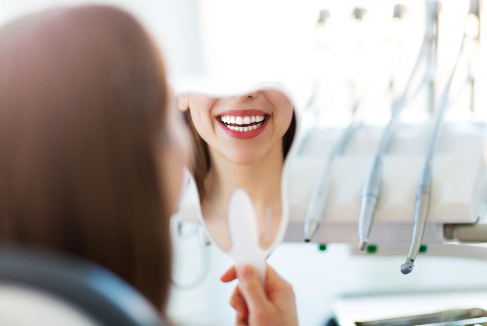 A woman smiles at her teeth in a dental mirror in a dentist's office.