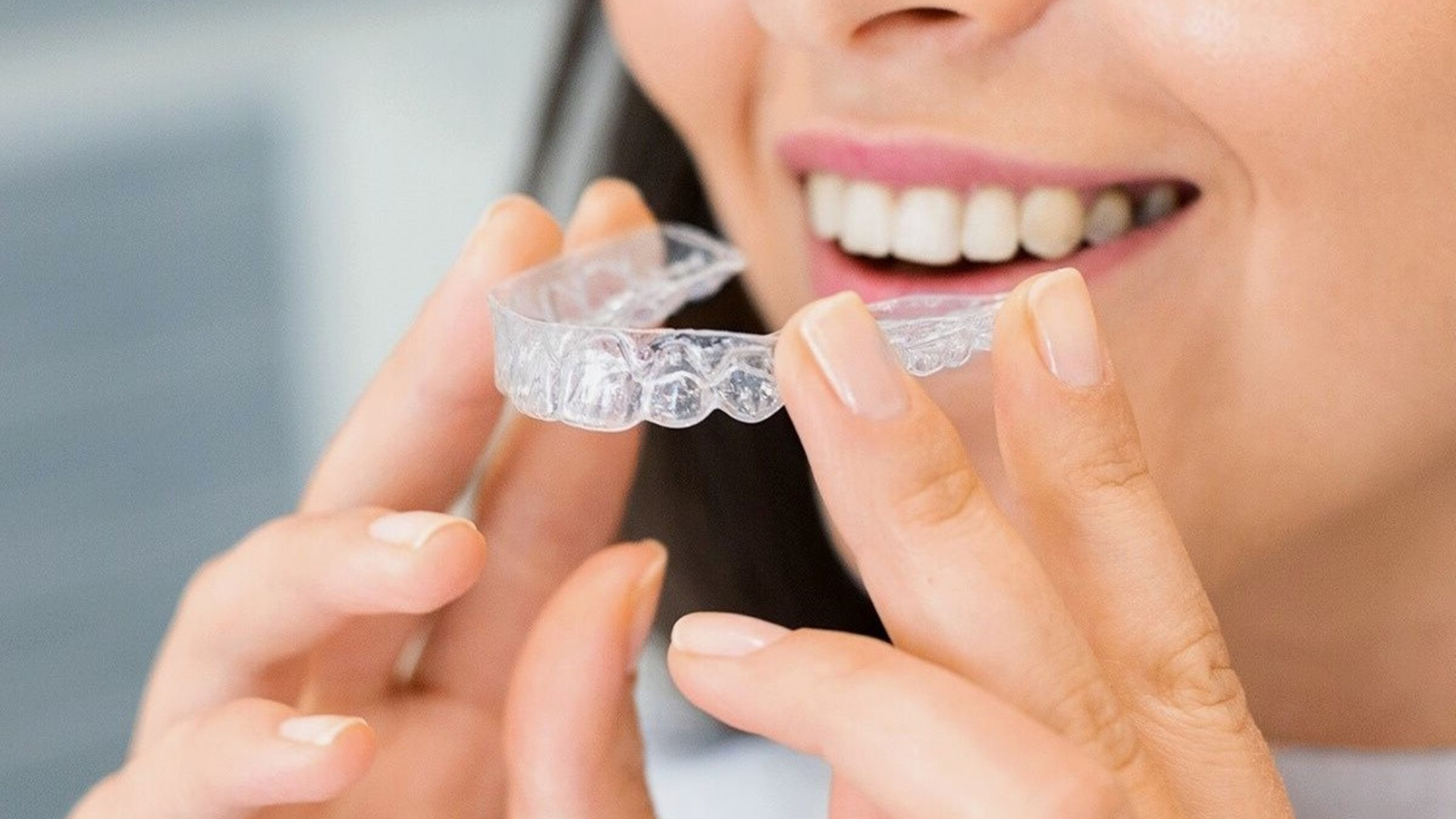 Woman smiling, holding clear dental aligner in front of teeth.