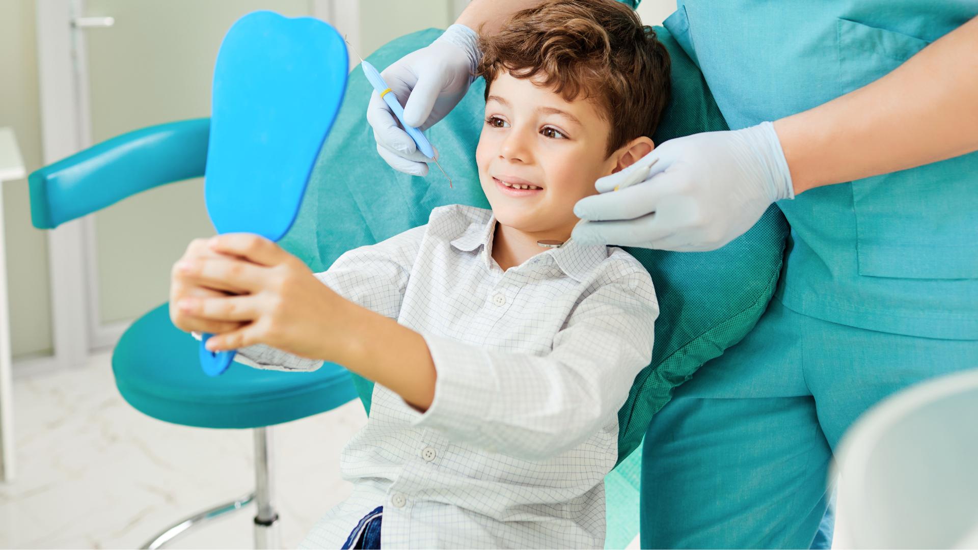 Boy in dentist chair looking at teeth in a mirror, dental assistant beside him.