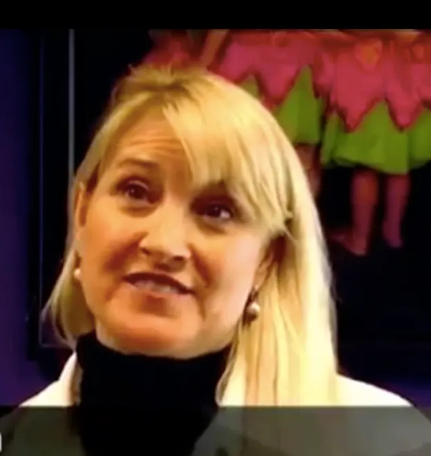 Woman with blonde hair wearing a black turtleneck, speaking. In the background, colorful doll costumes.