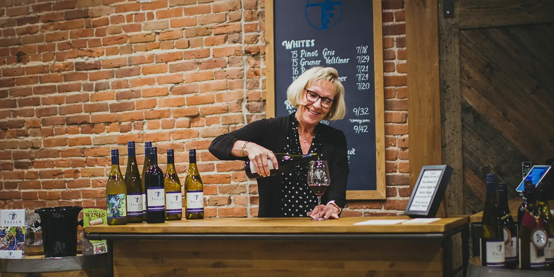 Woman pouring wine at a bar with bottles behind her. Brick wall in the background.