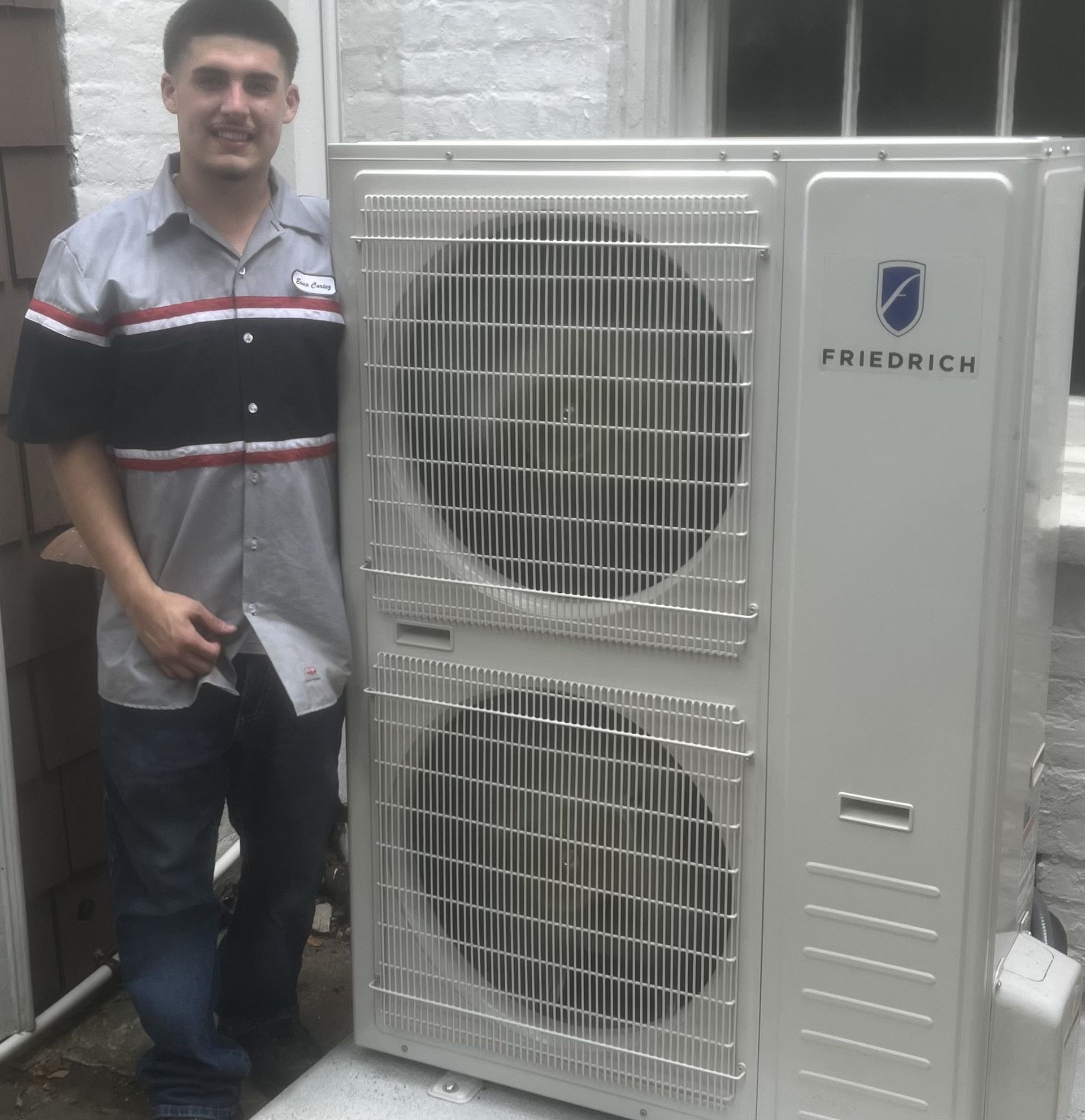 Man in work uniform stands next to a white Friedrich air conditioning unit outdoors.