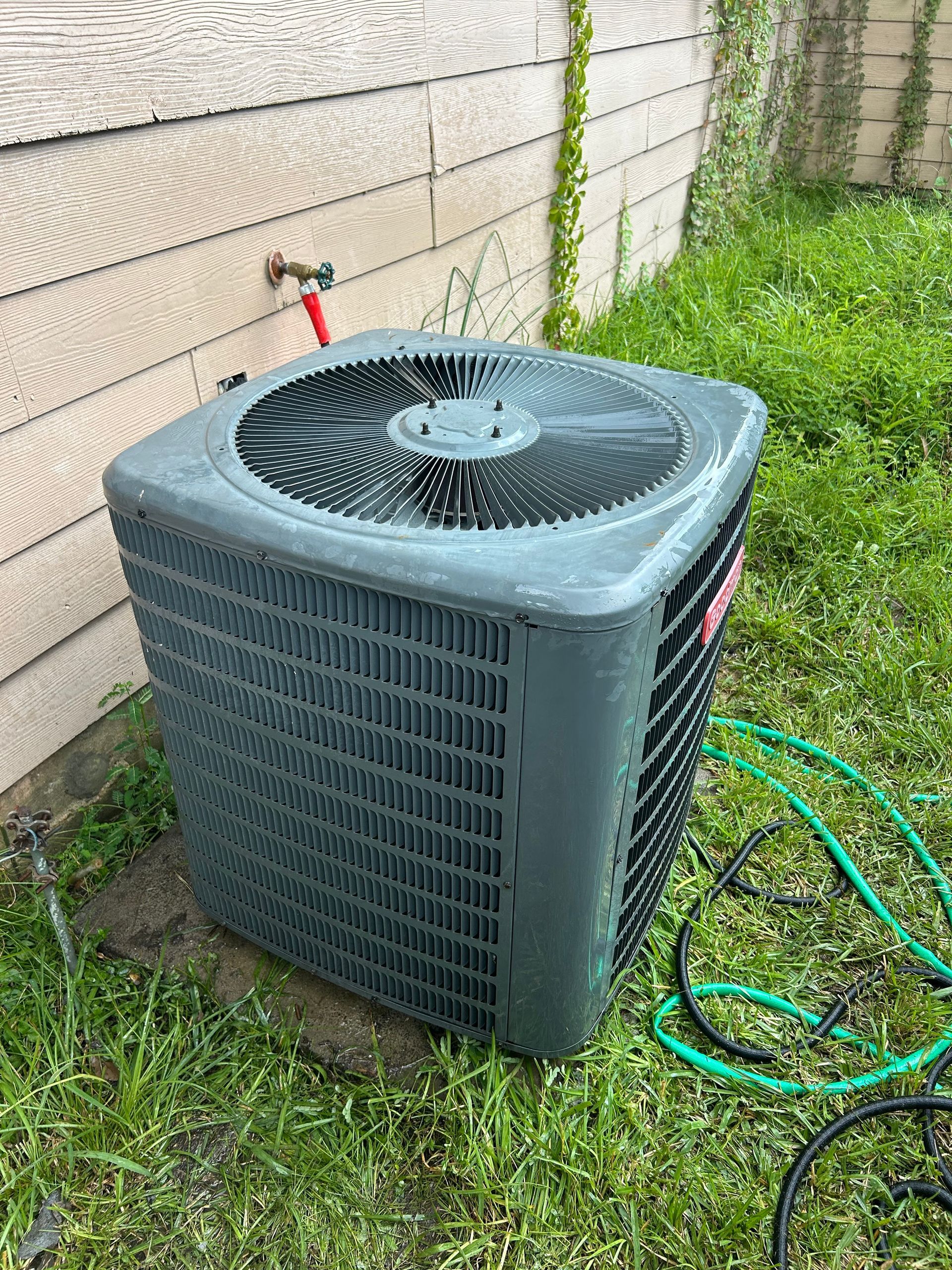 An air conditioning unit outside a building is being cleaned with soapy water. Green hose nearby.