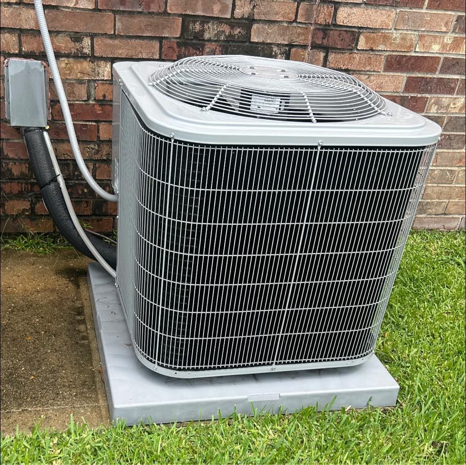 An air conditioning unit on a concrete pad near a brick wall and grass.