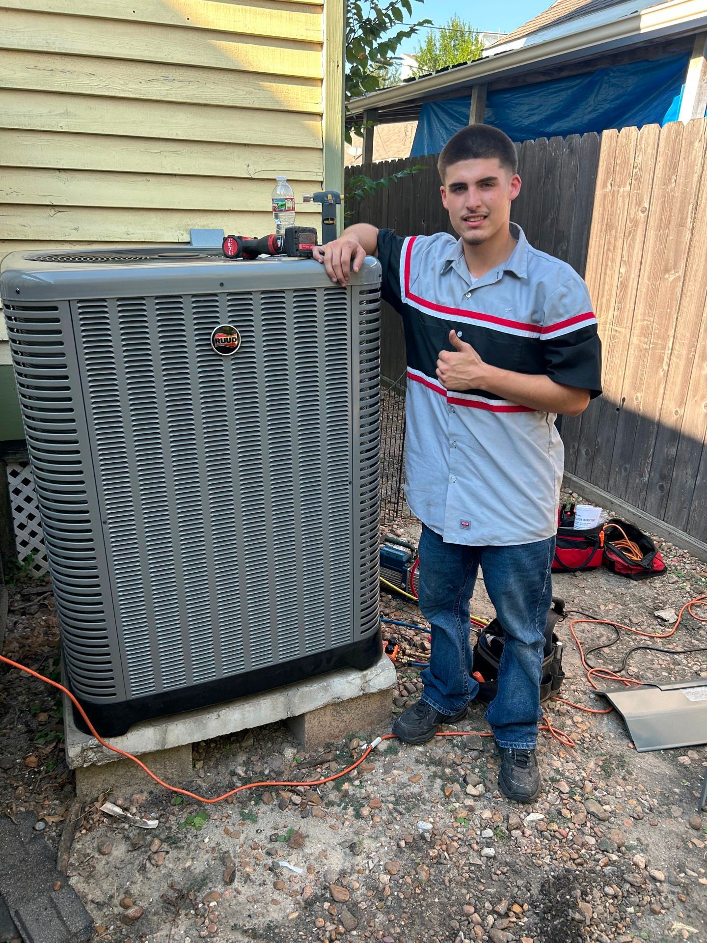 Man in work shirt stands next to an HVAC unit, giving a thumbs up.