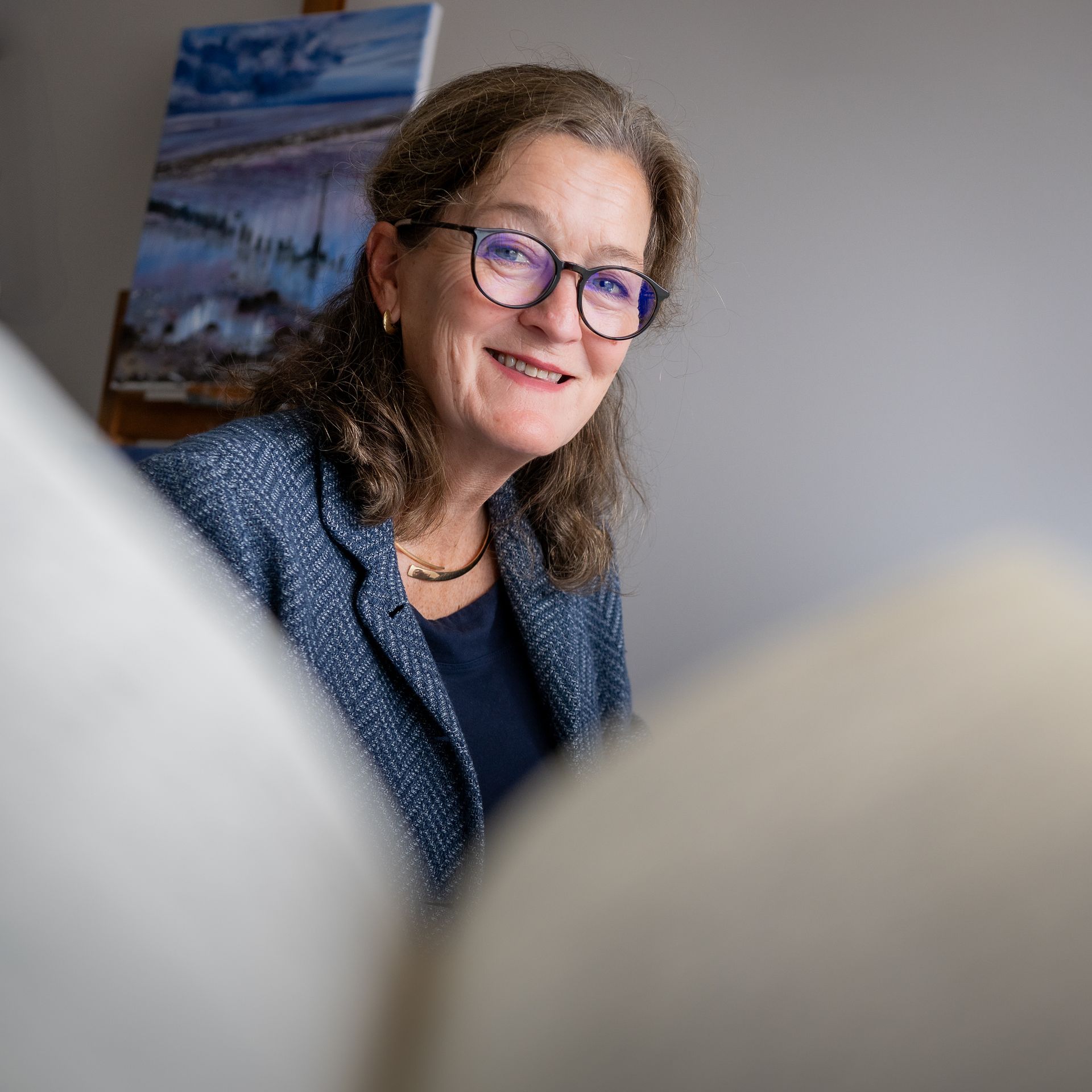 Een vrouw met een bril en een blauw jasje staat lachend voor een schilderij.