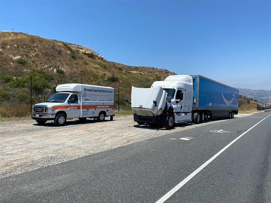 A white Amazon semi-truck with hood up beside an orange service van on a highway; hills in background.