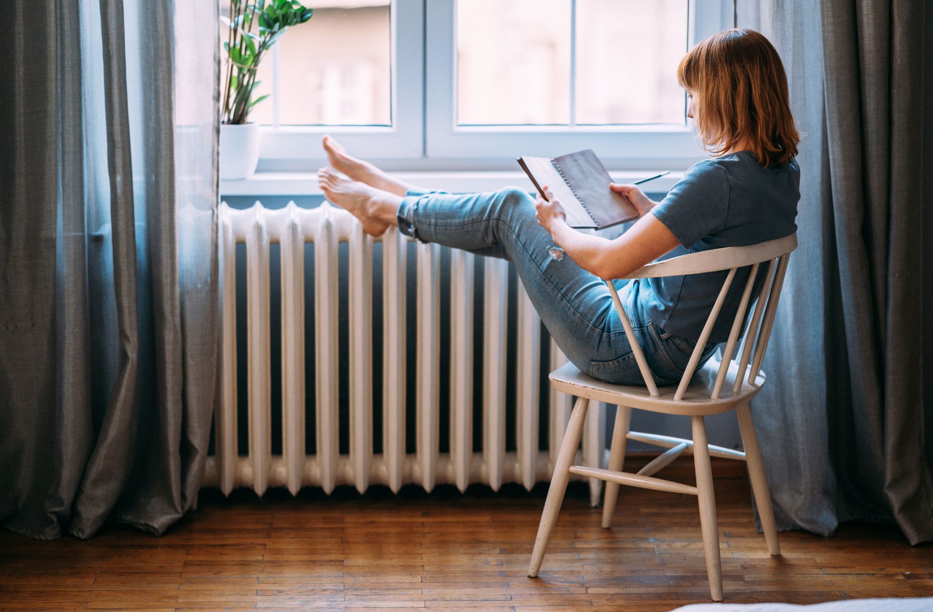 A woman is sitting in a chair in front of a radiator reading a book.