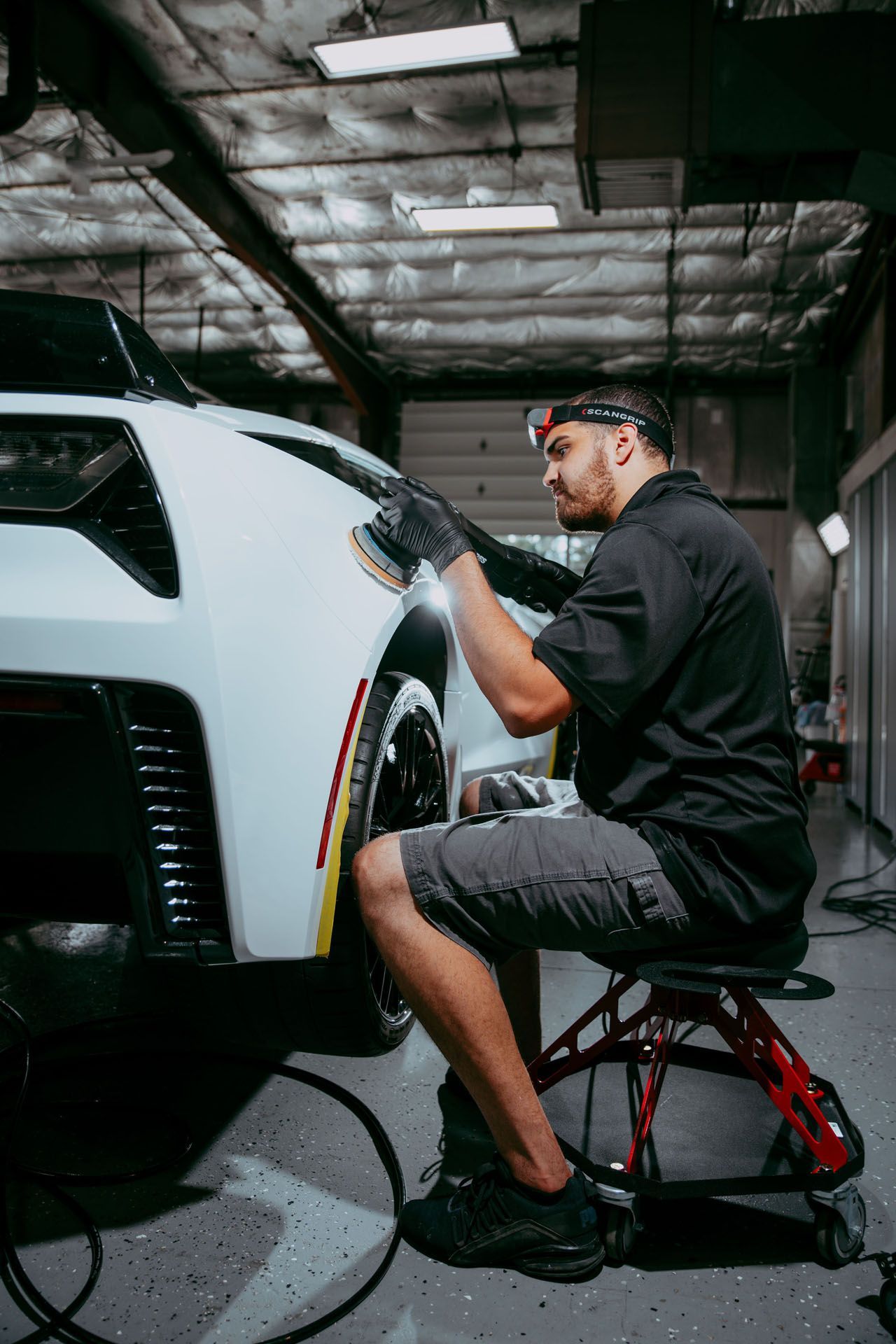 A man is sitting on a stool polishing a white car.