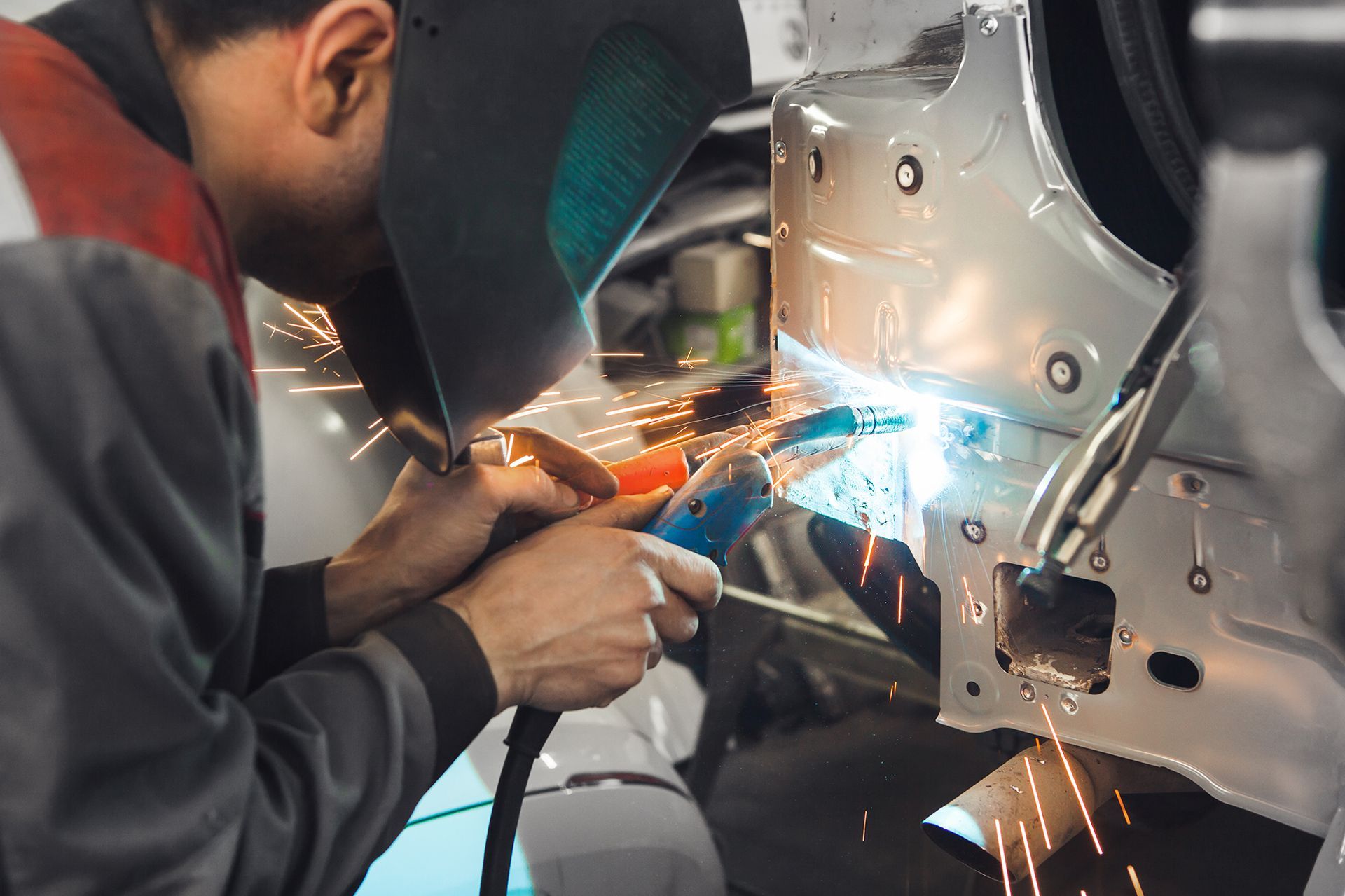 Welder in protective mask sparks while working on car body in auto shop.