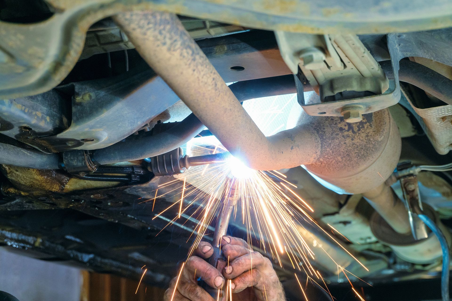 Hands welding an exhaust pipe on the underside of a car, sparks flying in a garage setting.