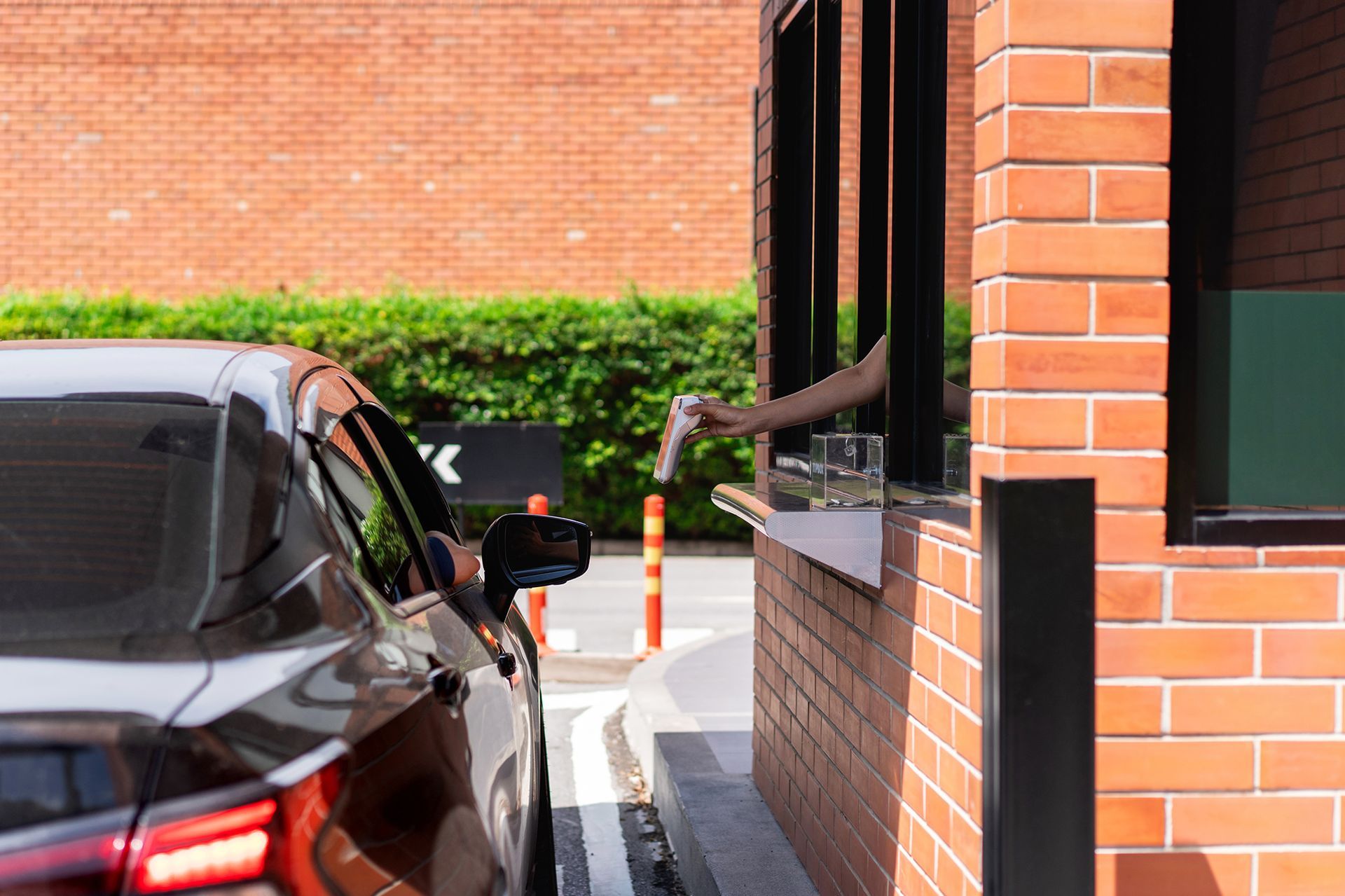 A person's arm handing a drink through a drive-thru window to a driver in a black car at a brick building.