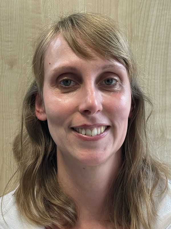 Frau mit heller Haut und blondem Haar lächelt vor einem hellen Holzmaserungshintergrund in die Kamera.