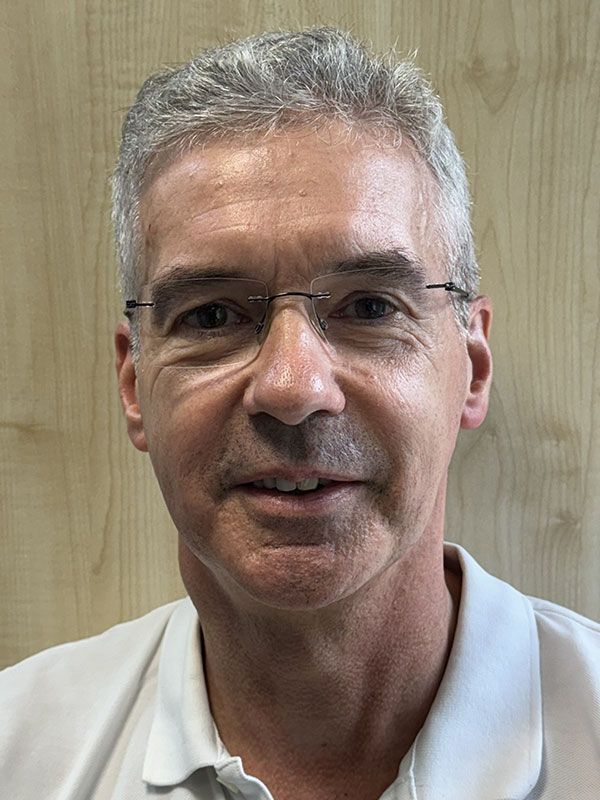 Mann mit ergrauendem Haar, Brille, trägt ein weißes Poloshirt und blickt direkt in die Kamera.