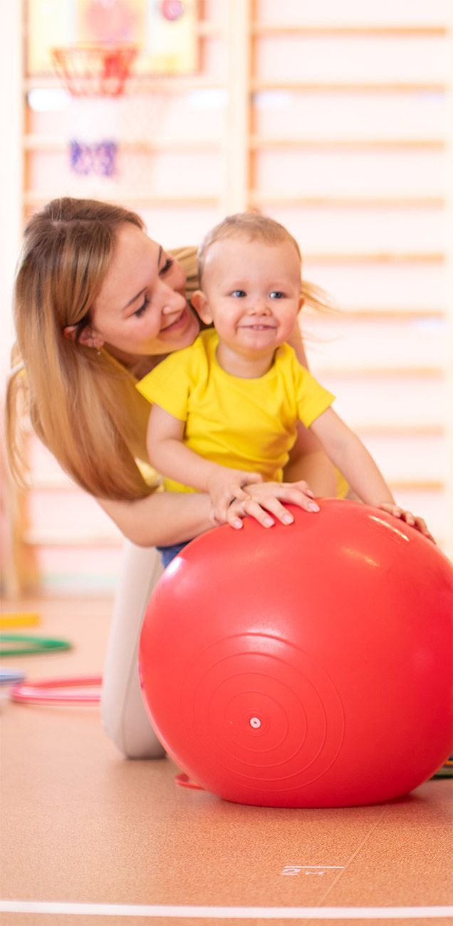 A woman and a baby are playing with a red exercise ball.