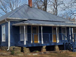 Blue house with porch and steps, stone foundation, metal roof, trees in background.