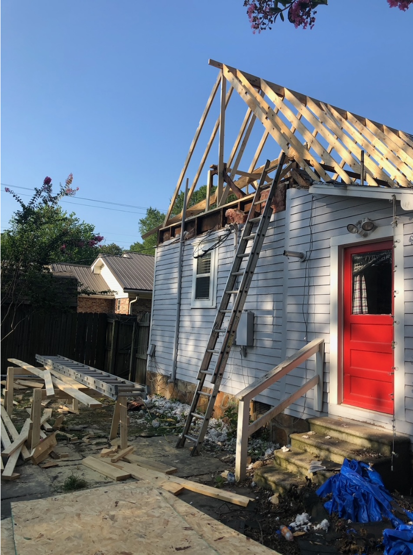 House under construction; wood frame roof, white siding, red door, ladder, blue tarp.