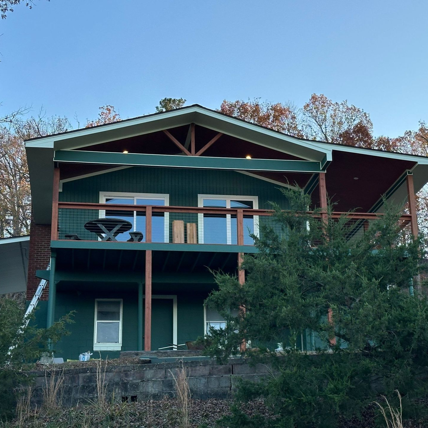 Green house with deck, windows, brown trim, and a red roof against a blue sky.