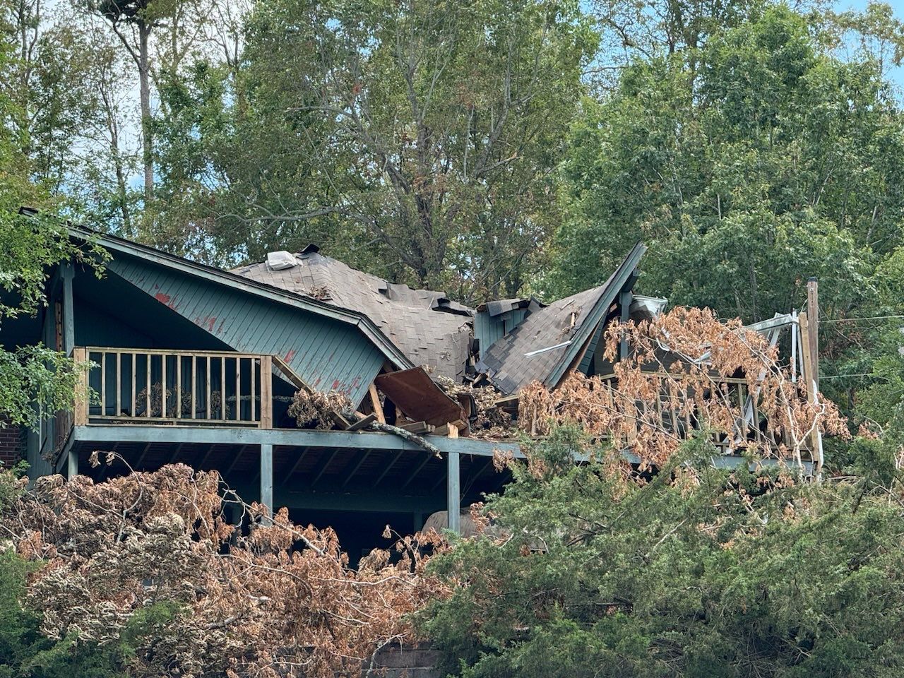 A damaged house with a collapsed roof, surrounded by trees and foliage.