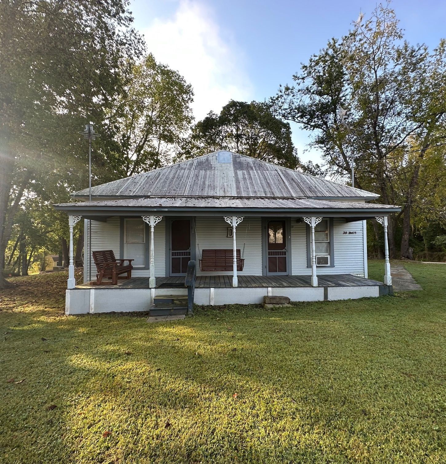 A weathered, white one-story house with a porch, surrounded by trees and green grass.