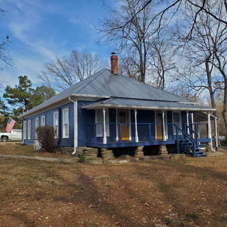 Blue house with porch, raised foundation, blue roof, and a red brick chimney against a blue sky.