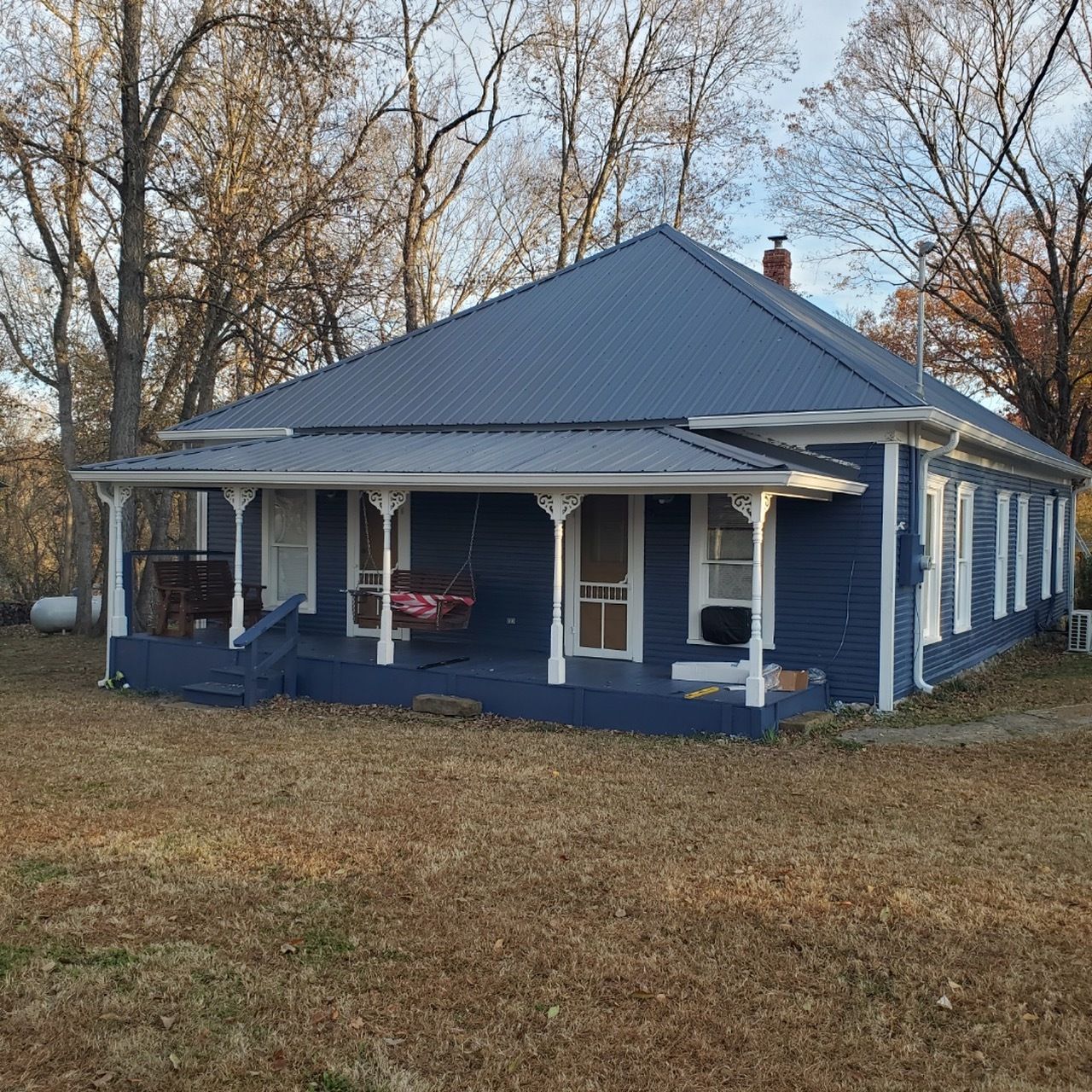 Blue-painted house with porch and metal roof on a grassy lot surrounded by trees.