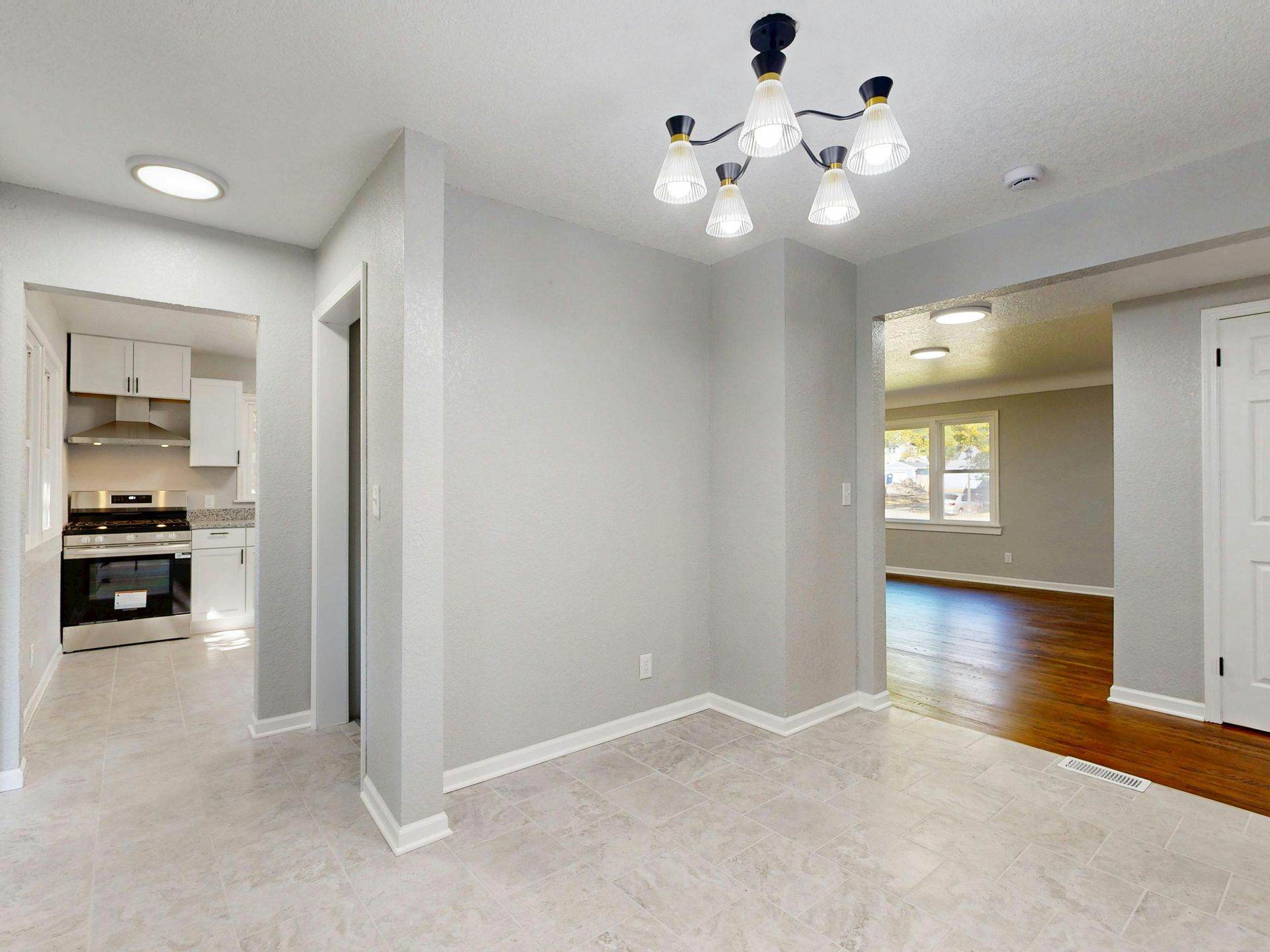 Empty modern dining room with gray walls, white carpet, chandelier, and a view into the kitchen and hardwood floor.