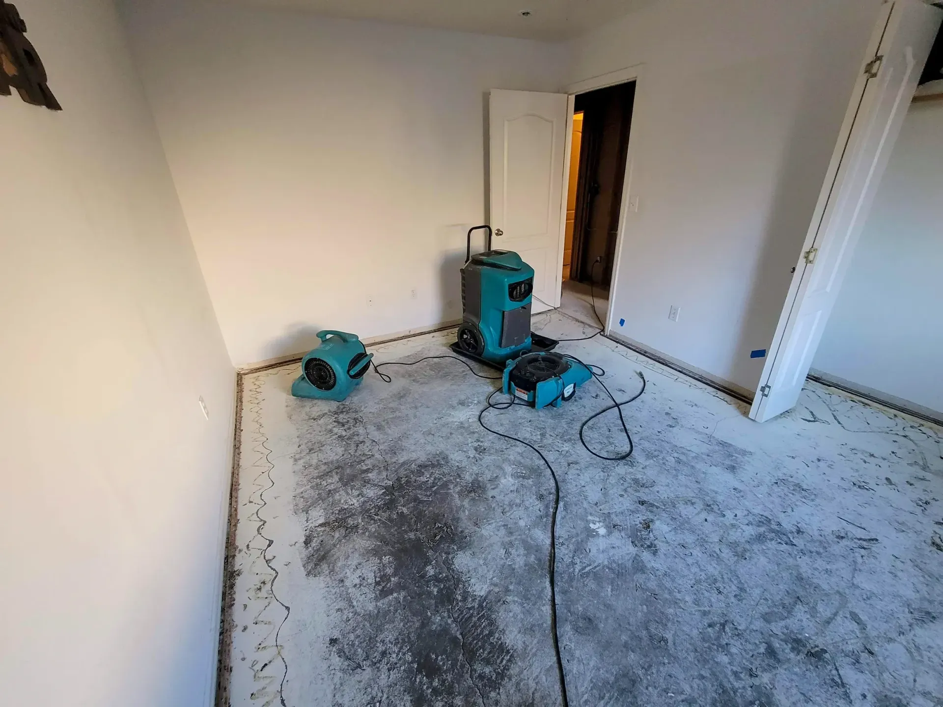 A room undergoing water damage restoration, featuring teal industrial dehumidifiers and air movers on a bare subfloor.