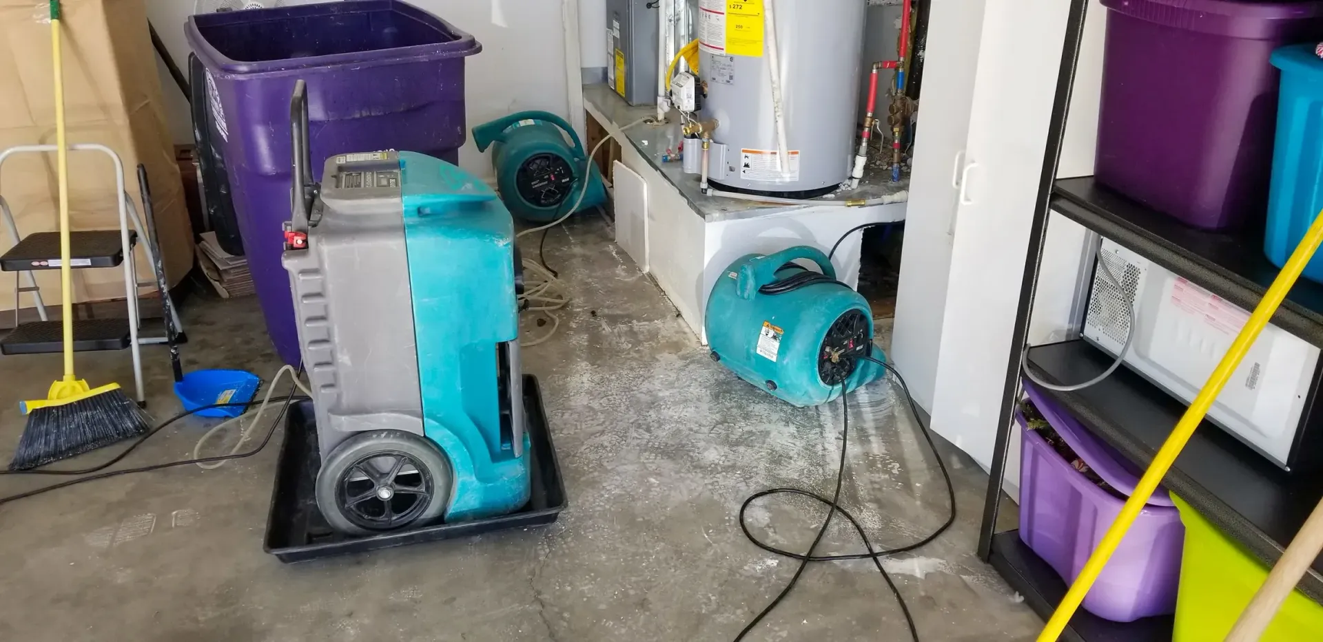 Industrial dehumidifiers and floor fans dry a water-damaged garage floor near a water heater.