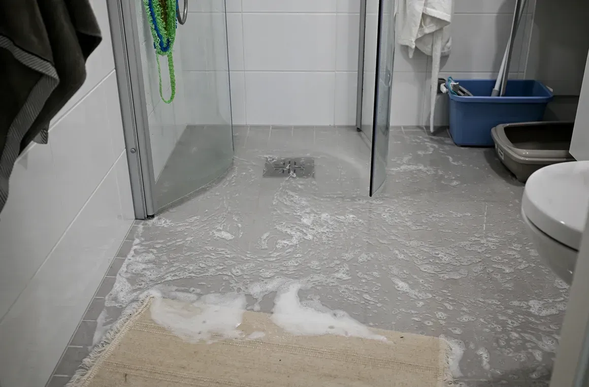 A bathroom floor is covered with white foam and residue, leading toward a glass shower enclosure with a drain.
