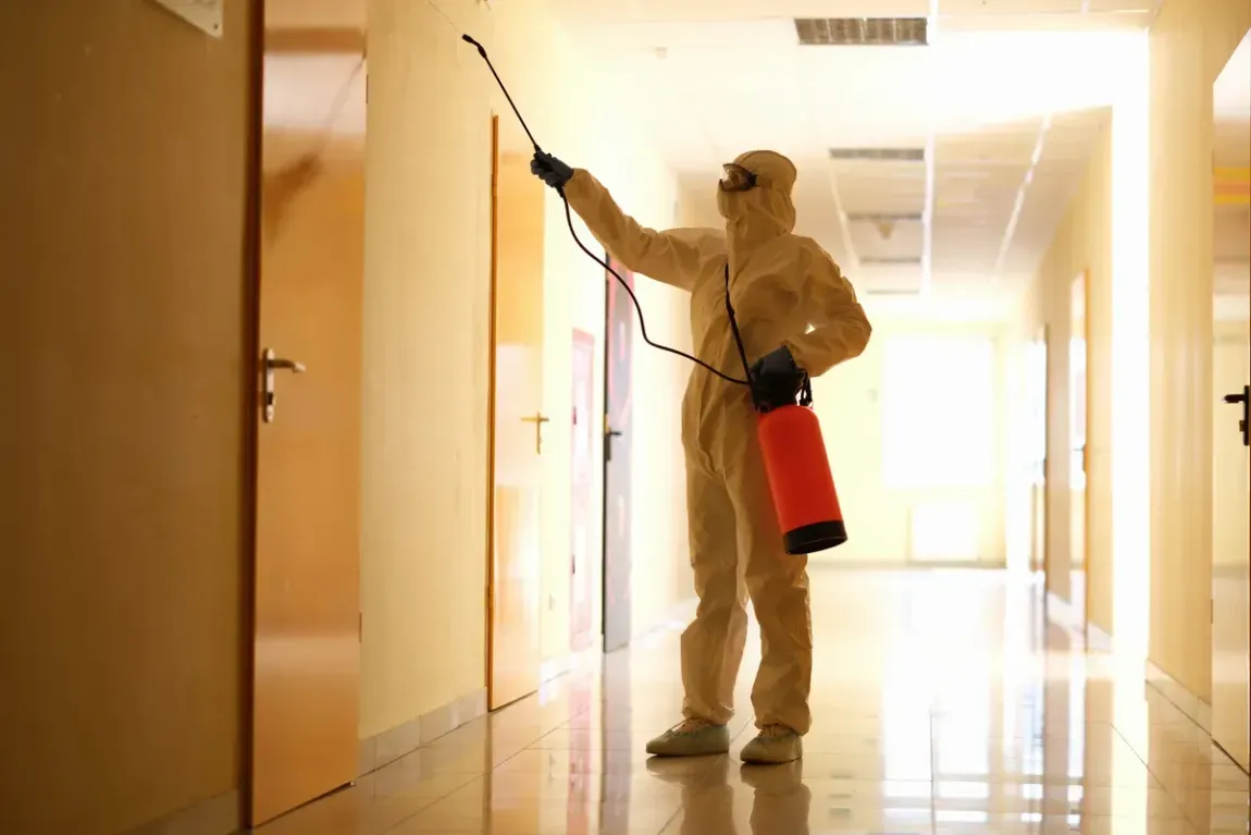 A person in a protective suit sprays disinfectant on a hallway wall in a brightly lit building.