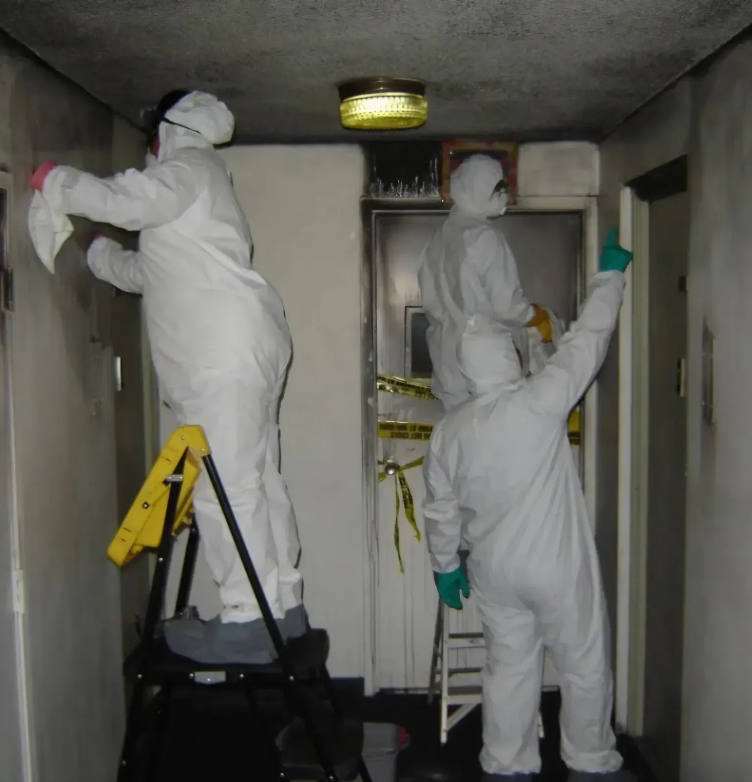 Workers in white coveralls painting a narrow room’s ceiling and walls with rollers and ladders.