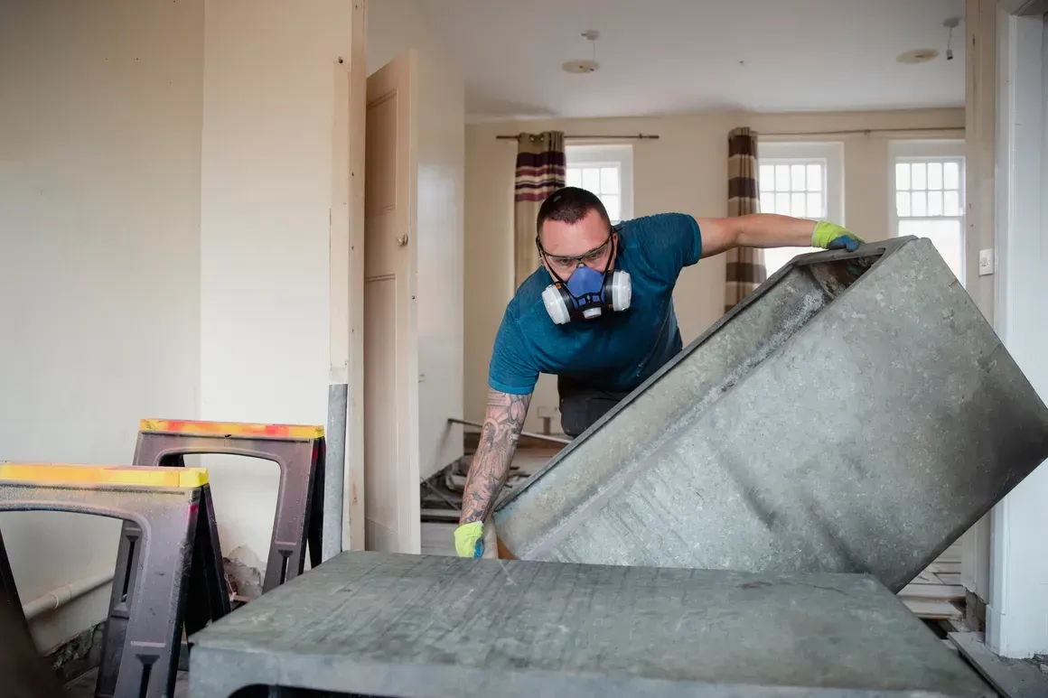 A person in a respirator and gloves lifts a heavy concrete slab inside a room undergoing renovation.