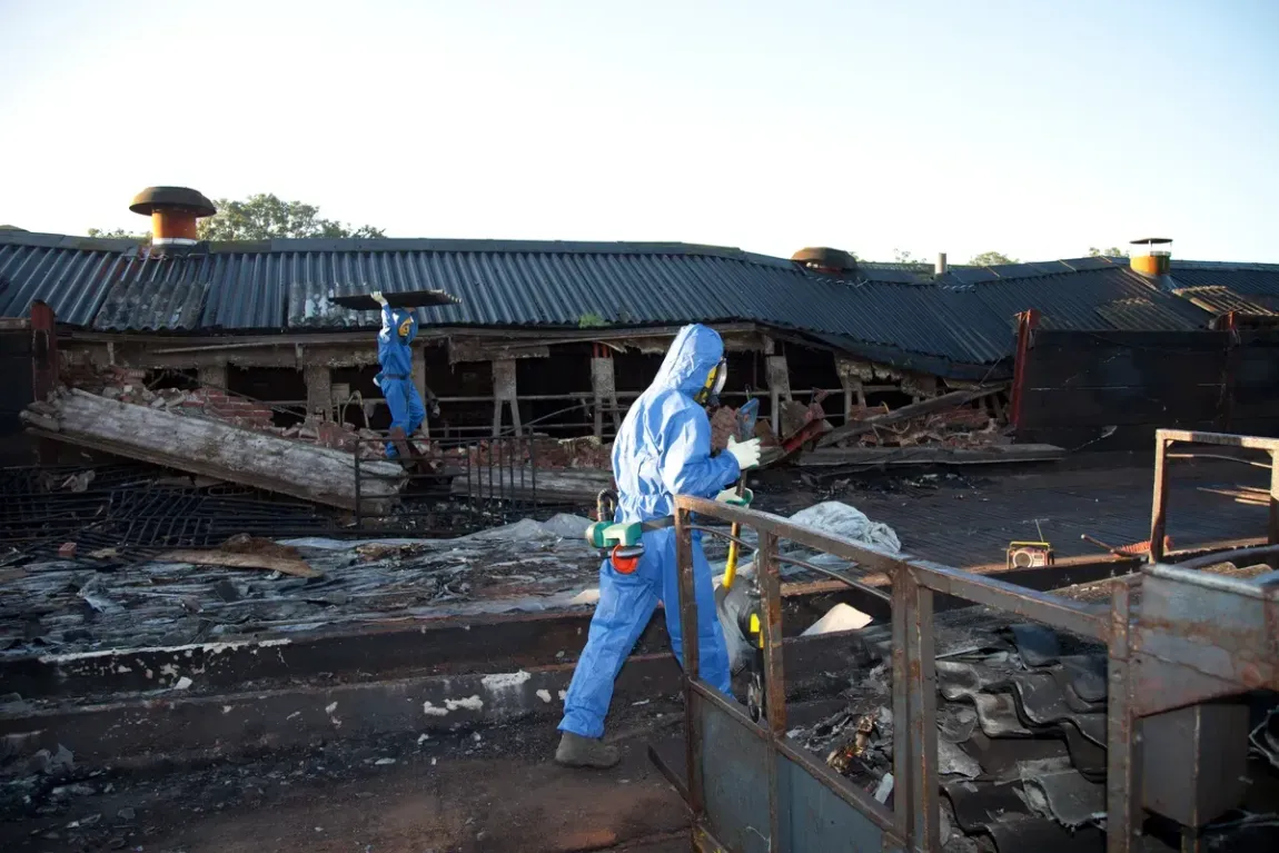 Two people in blue protective suits work amid the debris and charred remains of a destroyed building.