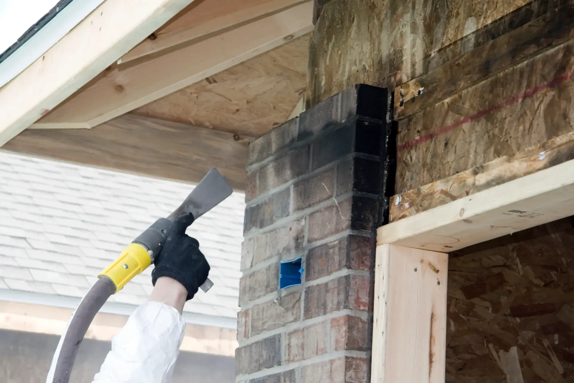 A gloved hand uses a spray tool to apply dark sealant to the edge of a brick wall on a construction site.
