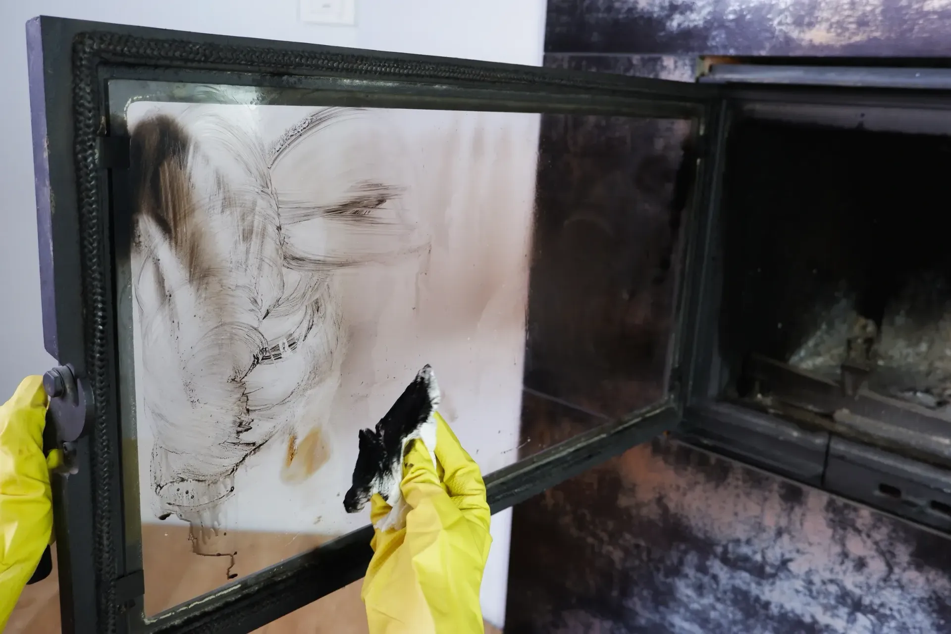 A person wearing yellow rubber gloves cleans soot and residue off a fireplace glass door with a cloth.
