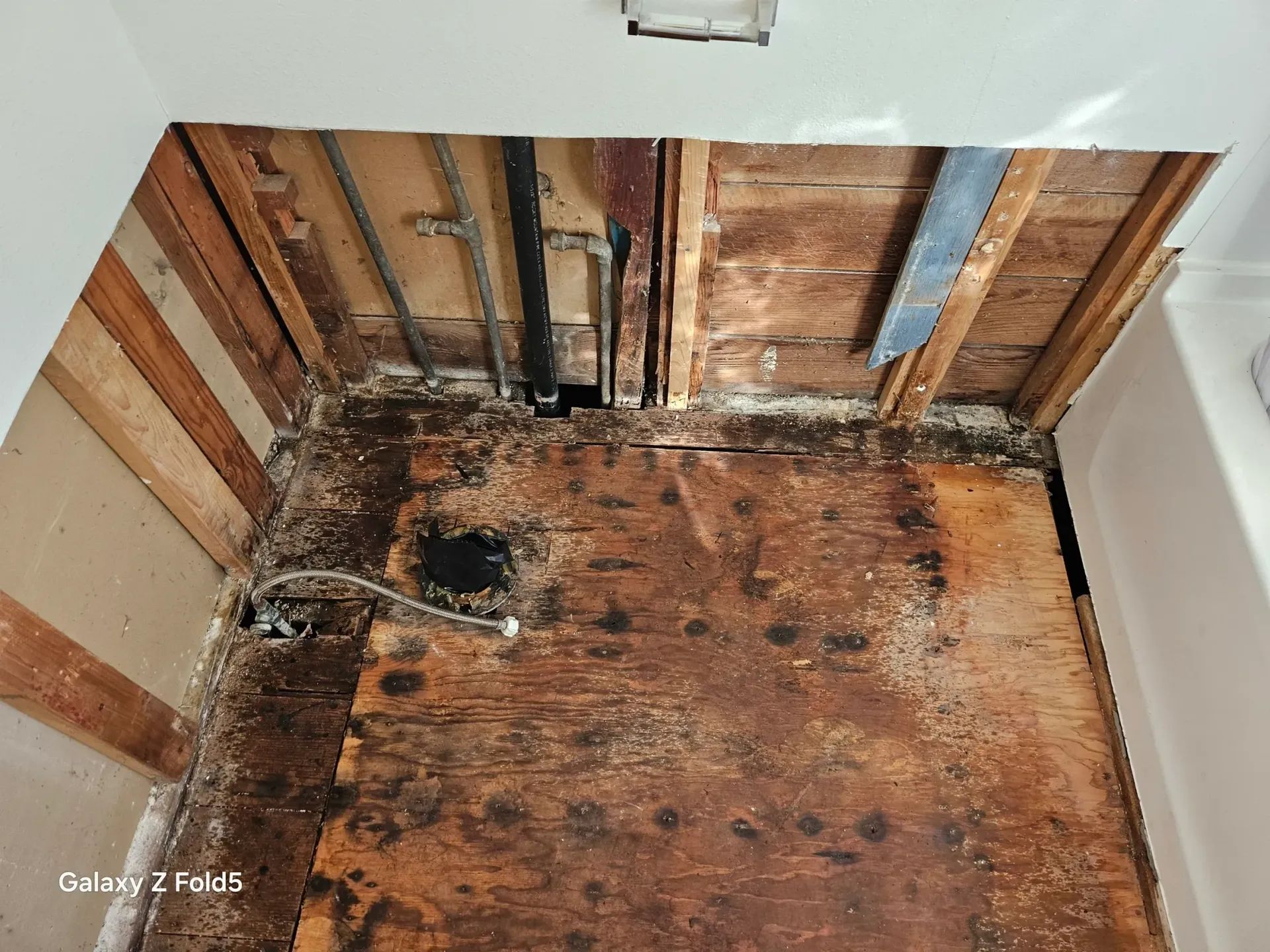 An open bathroom floor with severe water damage, dark rot, and exposed studs, next to a white bathtub.