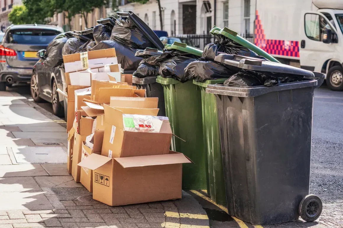 A row of black and green trash bins overflowing with bags on a city sidewalk next to a stack of cardboard boxes.