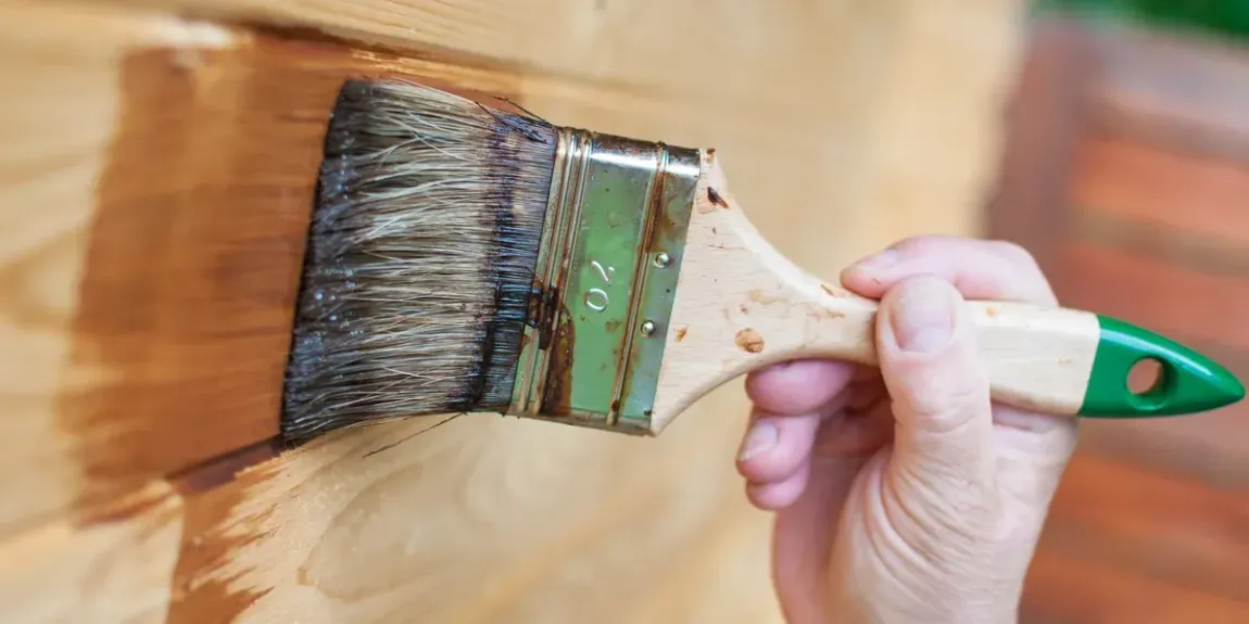 A hand holding a paintbrush applies brown wood stain to a light-colored wooden surface.
