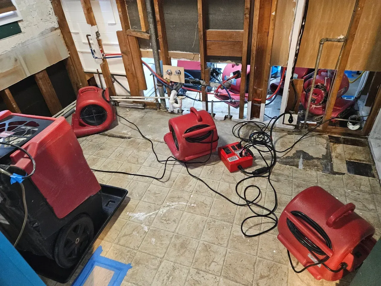 Industrial dehumidifiers and red air movers dry a water-damaged room with exposed wall studs and open plumbing.