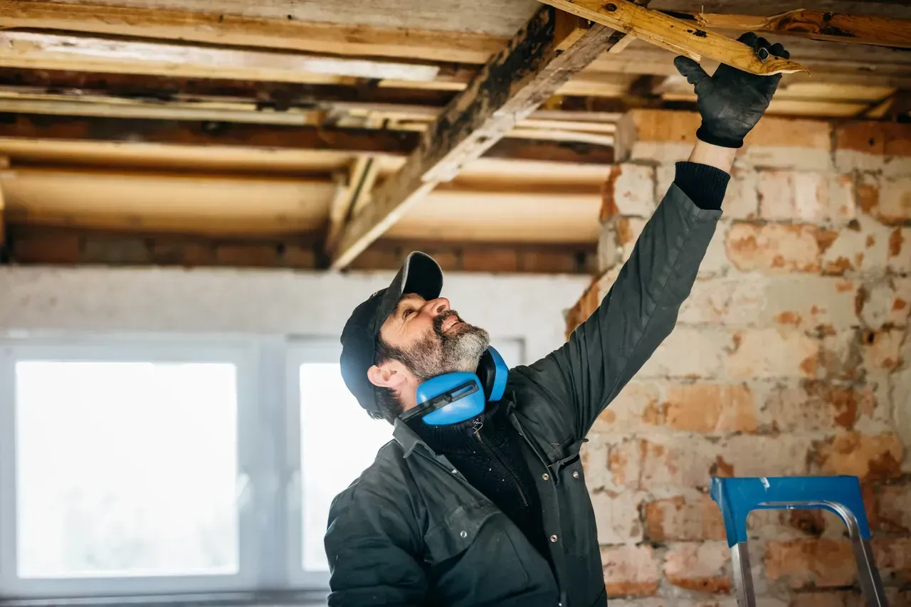 A person in a work jacket, cap, and ear protection examining a wooden ceiling beam inside an unfinished brick building.